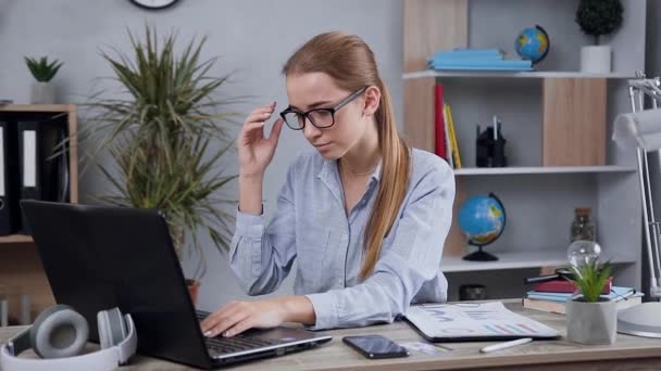 Крупним планом втомлена приємна 25-річна дівчина, яка працює за комп'ютером, знімає окуляри, розтирає очі і продовжує працювати — стокове відео