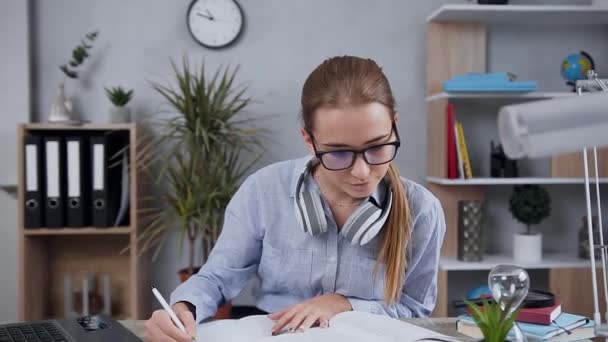 Koncentrerad blond attraktiv flicka i glasögon sitter vid bordet och skriver uppsats med hjälp av bok — Stockvideo