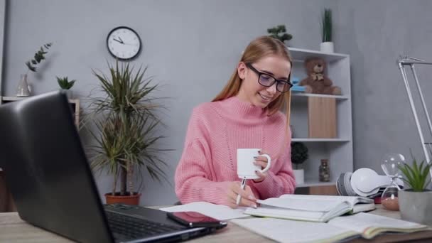 Pembe kazaklı neşeli sarışın kız kitaptan deftere gerekli notları yazıyor ve çay içiyor. — Stok video