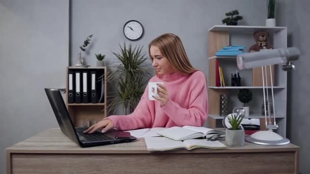 Mouvement lent d'agréable jeune fille heureuse en pull tricoté qui pose sur la caméra avec une tasse de thé pendant le travail à l'ordinateur — Video