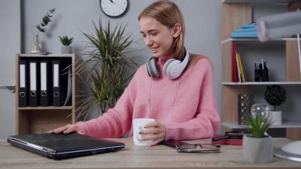 Adorável jovem feliz vestida com camisola de malha que caminha para a mesa, abrindo o laptop, colocando fones de ouvido e desfrutando de música bonita — Vídeo de Stock