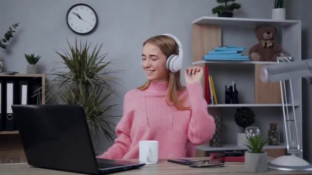 Sonriente jovencita guapa con auriculares vestidos con suéter de punto rosa sentada a la mesa y haciendo movimientos de baile cuando escuchas música moderna — Vídeo de stock