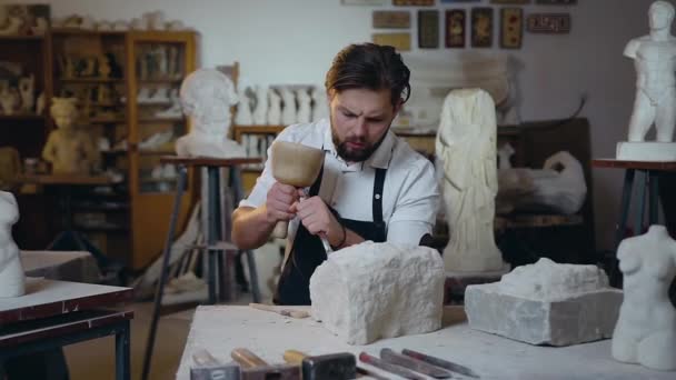 Aantrekkelijke 30-jarige bebaarde meester zittend in de ambachtelijke studio en het afslaan van kalksteen de toekomstige sculptuur met behulp van hamer en beitel en later blazen de overblijfselen van kalksteen — Stockvideo