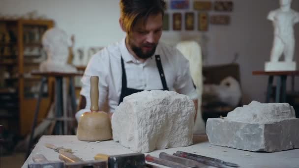 Knappe creatieve bebaarde jonge beeldhouwwerk caming naar de werkplek en zetten op glazen beginnen te werken met witte kalksteen met behulp van hamer en beitel — Stockvideo