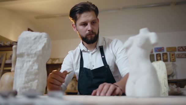 Hermoso joven escultor barbudo attantive mirando la escultura original haciendo marcas en la escultura futura en su lugar de trabajo — Vídeos de Stock