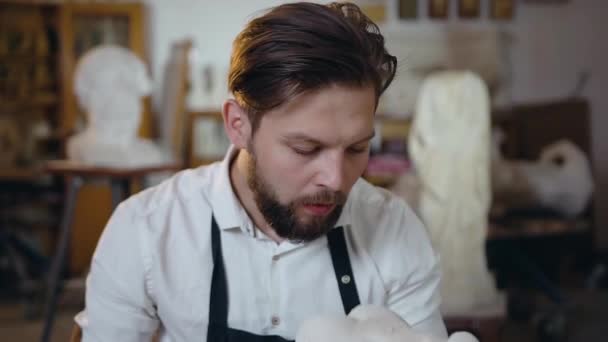 Slow motion of appealing attantive bearded sculptor in white shirt and apron which holding in his hands sculpture and blowing out from it the remains of limestone — 비디오