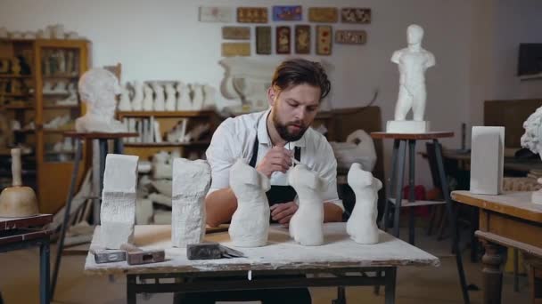 Front view of handsome talanted 30-aged male sculptor with beard which sitting at his workplace and scraping the remains of limestone with a brush from sculptures — 비디오