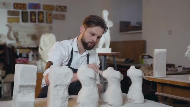 Aantrekkelijk portret van een ervaren, plezierige bebaarde beeldhouwer die zorgvuldig de resten van kalksteen van handgemaakte sculpturen schoonmaakt — Stockvideo