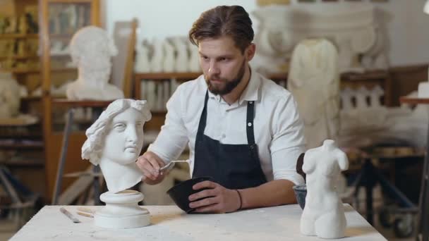 Привлекательный молодой современный бородатый скульптор аккуратно накладывает клей на голову скульптуры — стоковое видео