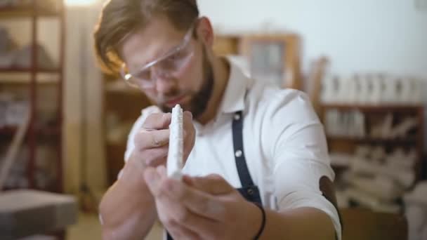 Boa aparência cuidado jovem barbudo mestre com corte de cabelo moda vestindo óculos especiais verificando o detalhe para a escultura — Vídeo de Stock