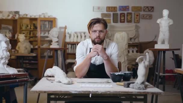 Vooraanzicht van aangename attantieve moderne beeldhouwer met baard in bril zittend op zijn werkplek in de studio en poserend op camera met oprechte glimlach — Stockvideo