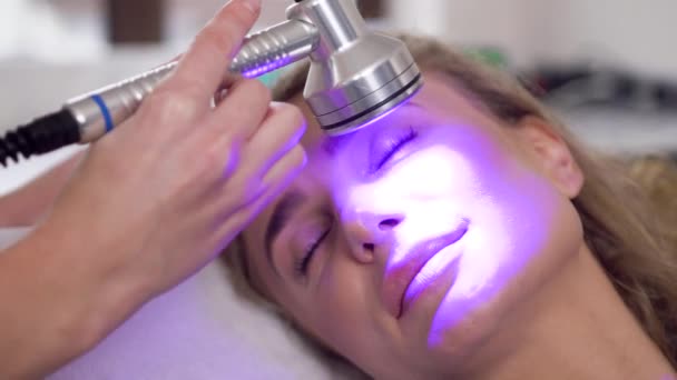 Primer plano de la tranquila mujer rubia atractiva que hace el rejuvenecimiento de su cara utilizando la herramienta ultrasónica especial en el salón de spa — Vídeos de Stock