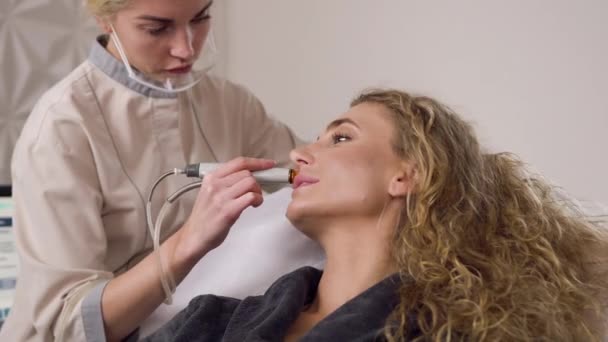 Cosmetologist feminino concentrado bonito que faz a limpeza ultrasônica no rosto de mulheres no salão de beleza — Vídeo de Stock