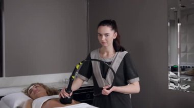 Gülümseyen profesyonel kadın masör kadınların karnında rf-lift ameliyatı yapıyor ve kamera önünde poz veriyor.