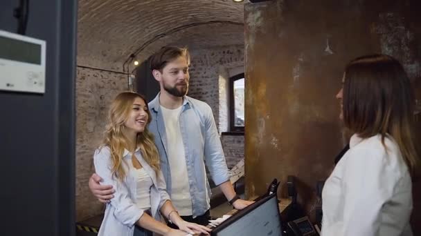 Good-looking modern bearded guy hugging his charming happy girl while talking with friendly female recepcionist at the desk in hotel — Stock Video