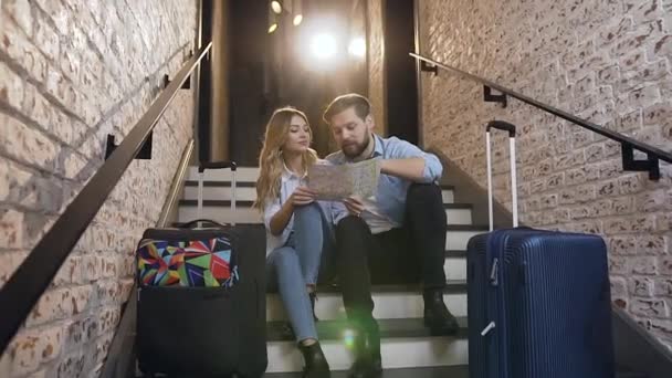 Aantrekkelijk portret van opgetogen charmant jong stel dat in het modehotel op de trap zit en op zoek is naar de plattegrond van hun vakantie — Stockvideo