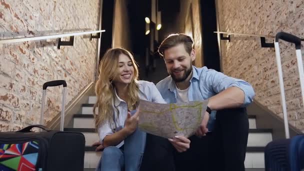 Lachend vrolijke jonge moderne man en vrouw zittend op een trap in een hedendaags hotel en vrolijk met elkaar naar de kaart kijkend — Stockvideo