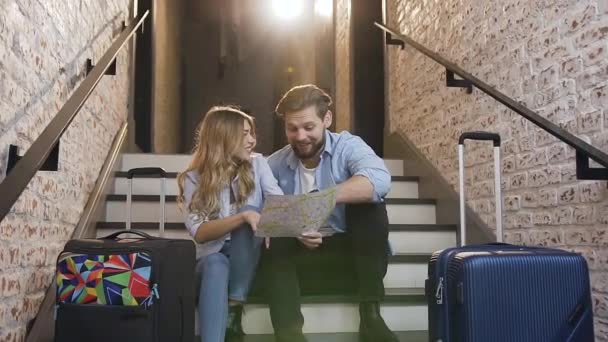 Slow motion of lovely cheerful young bearded guy with his blond girlfriend which sitting on the steps in hotel and looking at map talking about their vacation — 비디오