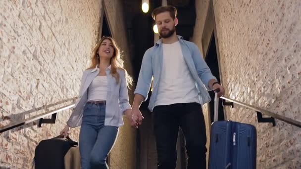 Счастливая прекрасная счастливая молодая пара с чемоданами спускается по лестнице в модный отель, держа за руки — стоковое видео