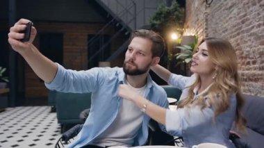 Appealing smiling blond woman hugging her joyful bearded boyfriend by neck during making funny selfie on the background of decorated brick wall