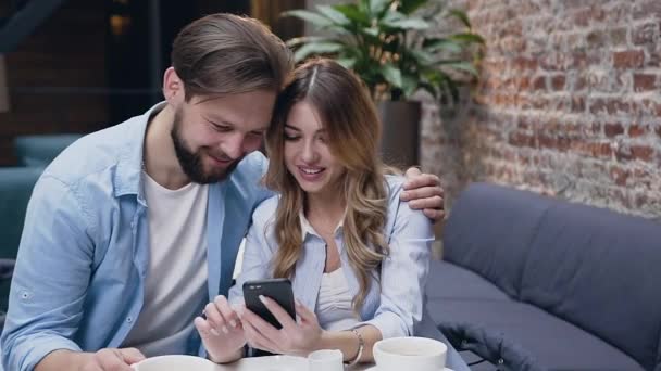 Весела сучасна стильна молода пара обіймає і дивиться за жіночим телефоном, сидячи за столом у готельному лобі — стокове відео