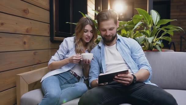 Cámara lenta de la encantadora y sonriente pareja moderna en el amor que utiliza la tableta PC mientras está sentado en el sofá suave en el vestíbulo del hotel — Vídeo de stock