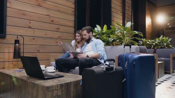 Přední pohled na sympatický romantický radostný pár, který sedí společně v hotelové hale a pomocí turistické mapy a tabletu PC hledat zajímavá místa k návštěvě — Stock video
