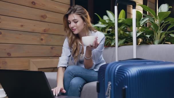 Mooie glimlachende jonge vrouw met een kopje thee en typen op de computer terwijl ze met haar bagage in de lobby van het hotel zit — Stockvideo