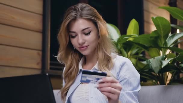 Atractivo retrato de una guapa joven rubia sonriente que ingresa los detalles de su tarjeta bancaria en el portátil mientras está sentada en el pasillo del hotel — Vídeo de stock