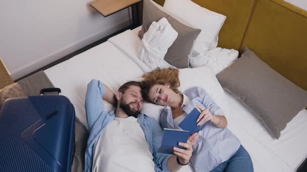 Вид с вершины романтической симпатичной влюбленной молодой пары, которая лежит на кровати отеля, пересматривая свои паспорта и смеясь — стоковое видео