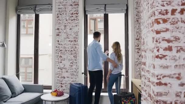 Обратный вид на влюбленную молодую счастливую современную пару, которая обнимает друг друга, глядя в большое окно в номере отеля — стоковое видео