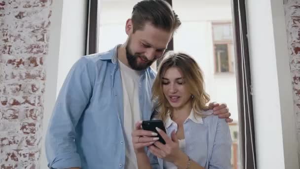 Vista da vicino di adorabile felice giovane coppia moderna innamorata che guardando il telefono delle ragazze e sorridendo da video o foto divertenti mentre in piedi vicino a una grande finestra — Video Stock