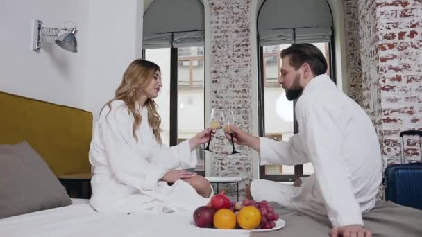 Adorable feliz pareja joven y amorosa en albornoces sentados en la cama y bebiendo champán en la habitación de hotel bellamente diseñada — Vídeos de Stock