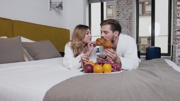 Вид спереди на привлекательную счастливую улыбающуюся молодую пару, одетую в халаты, которые лежат на кровати, а девушка кормит своего мальчика круассаном — стоковое видео