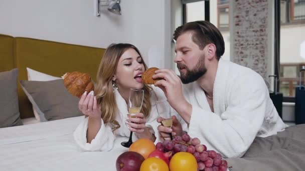 Front view of attractive cheerful bearded guy in bathrobe which feeding his smiling blond girlfriend with croissant and drinking together champagne while lying in hotel bed — 비디오