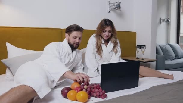 Conceito do hotel onde belo homem de barba sorridente em roupão de banho comendo uvas, deitado na cama, enquanto sua encantadora namorada loira feliz em roupão de banho terry usando laptop — Vídeo de Stock