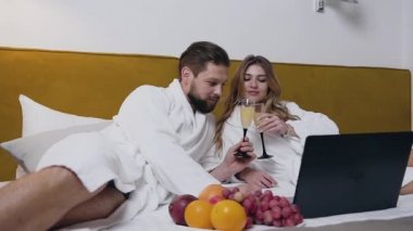 Beyaz bornozlu, otel odasında uzanıp köpüklü şarap içen mutlu, modern, şehvetli çiftin yakın görüntüsü.