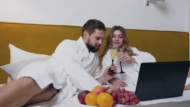 Vista próxima do jovem casal amoroso moderno feliz em roupões de banho brancos que deitado na cama e bebendo vinho espumante no quarto de hotel — Vídeo de Stock