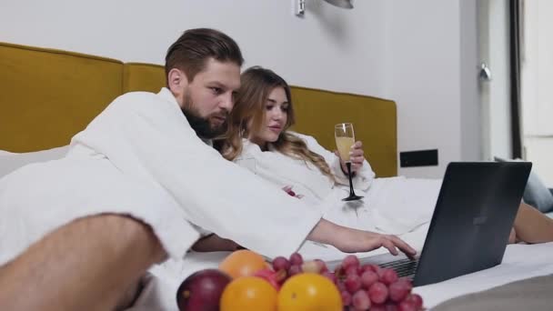 Vonzó amorált boldog fiatal pár fehér fürdőköpenyben fekszik a nagy ágyon a kortárs szállodában, és együtt nézik a számítógépen — Stock videók