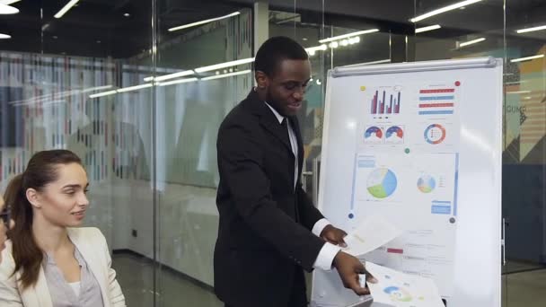 Atraente jovem empresário afro-americano transmitindo diferentes relatórios para seus parceiros de negócios bem-intencionados na sala de reuniões — Vídeo de Stock