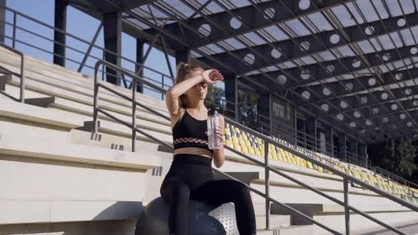 Довольно уставшая спортивная молодая женщина в спортивной одежде сидит на большом гимнастическом мяче и пьет воду во время спортивных тренировок на открытом воздухе — стоковое видео