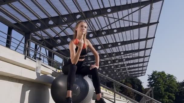 Front view of beautiful sporty modern young woman in fitness clothes which sitting on big ball and doing exercises with dumbbells outdoors — Stockvideo