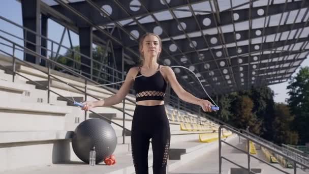 Zadowolona wysportowana, szczupła młoda kobieta w stroju fitness wykonująca ćwiczenia fizjologiczne ze skakanką podczas treningu sportowego na podium na stadionie — Wideo stockowe