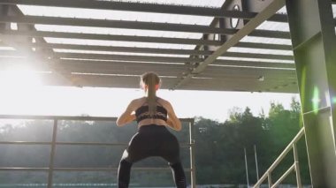 Back view of sexy slender young woman in fitness clothes which doing squat exercises outdoors under roof