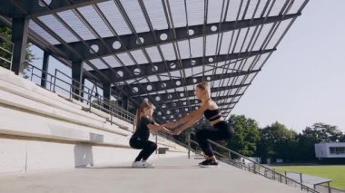 Sport training concept where two attractive sporty girls-elder and younger holding by hands and squating outdoors near stadium