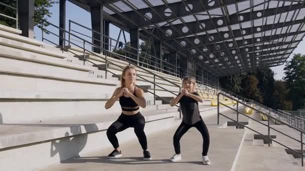 Vooraanzicht van aantrekkelijke atletische jonge vrouw en haar kind bedelaar die squat oefeningen doen op het podium van het stadion — Stockvideo