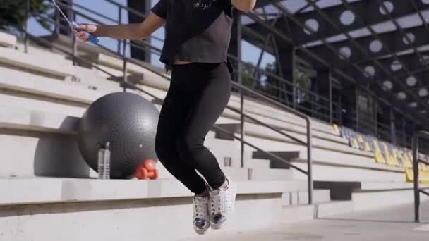 Aantrekkelijk portret van een lachend sportief klein meisje in zwarte sportkleding die springt met springtouw op het perron van het stadion — Stockvideo