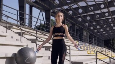 Front view of lovely slender sporty young woman in fitness clothes which doing jumping exercises with skipping rope on the tribune on the stadium