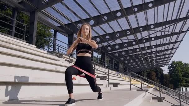 Приваблива струнка спортивна молода жінка з хвостом у чорному фітнес-одязі робить розтяжки з еластичною спеціальною стрічкою на відкритому повітрі — стокове відео