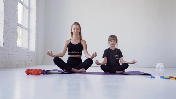 Attractive sporty calm two girls-elder and younger sitting on violet mats in lotus pose with closed eyes during yoga lesson — ストック動画